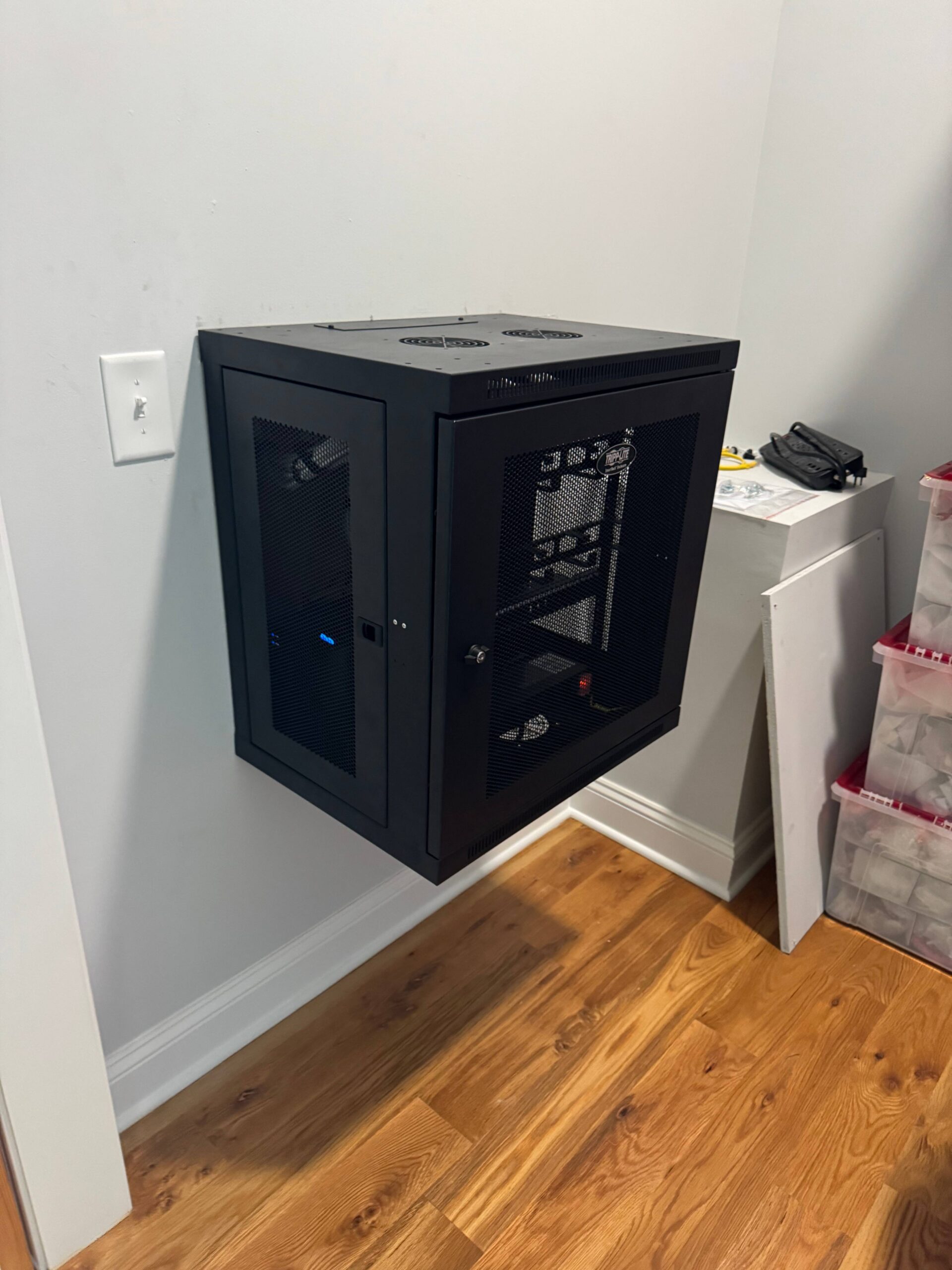Black wall-mounted server rack with glass front door, containing network equipment, is installed in a room with light walls and wood flooring.