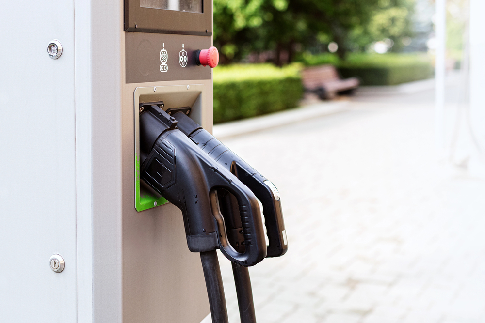 Close-up of electric vehicle charging station with two charging connectors plugged into the unit, positioned outdoors near a park bench and greenery.