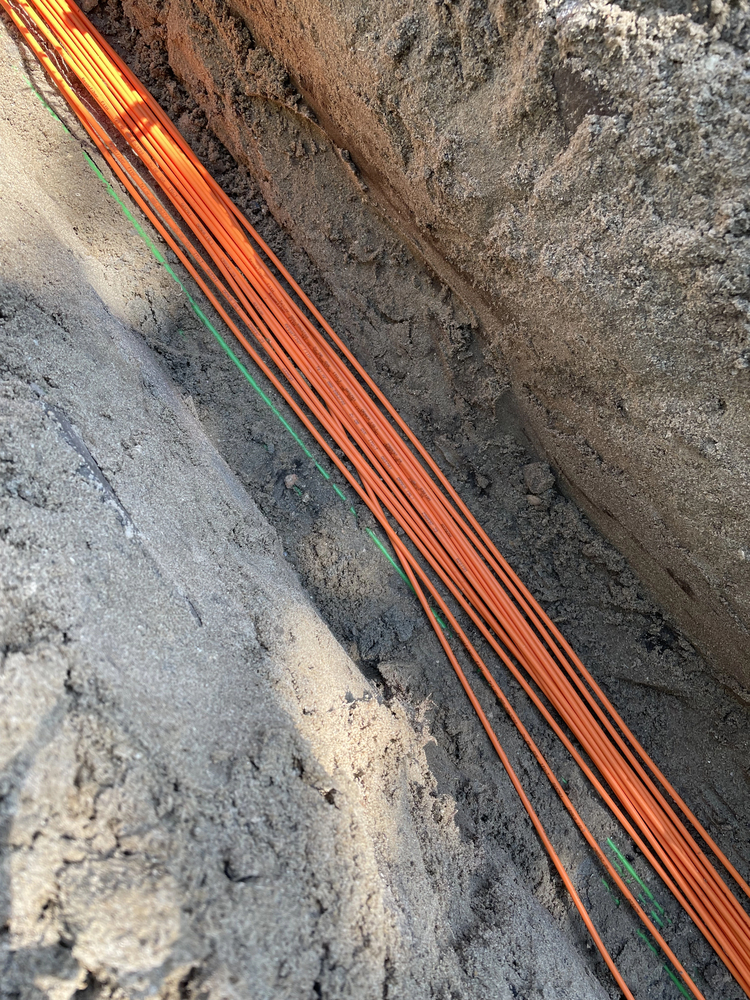 Several orange cables are laid in a trench in the ground with soil on both sides, likely as part of an underground utility installation.