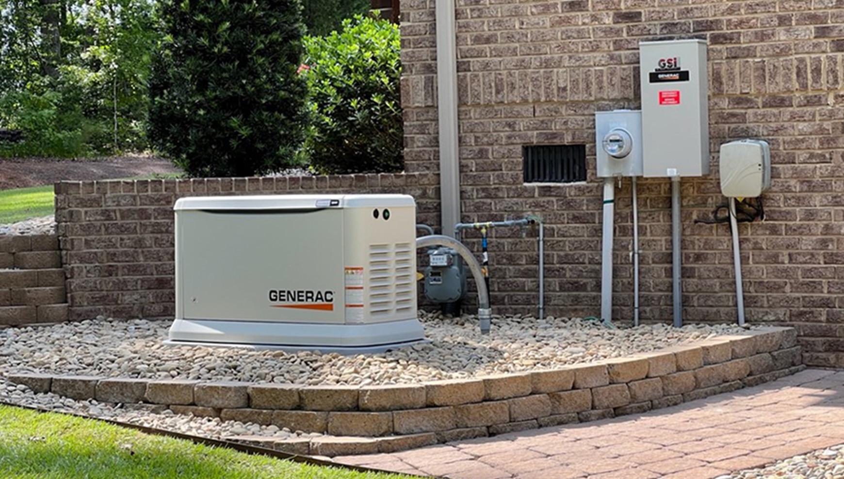 A Generac standby generator installed on a gravel bed next to a brick house, with utility meters and an electrical panel mounted on the exterior wall.