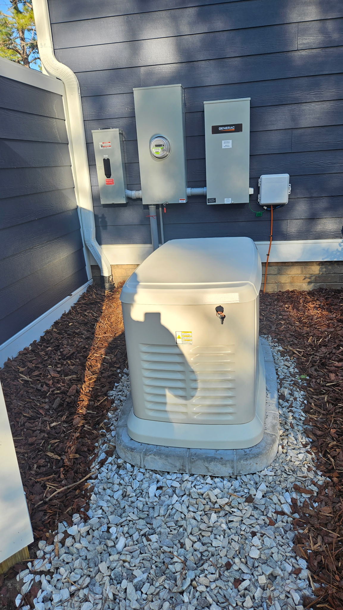 A standby generator is installed on a gravel pad outside a house, with electrical panels and utility meter mounted on the blue exterior wall behind it.