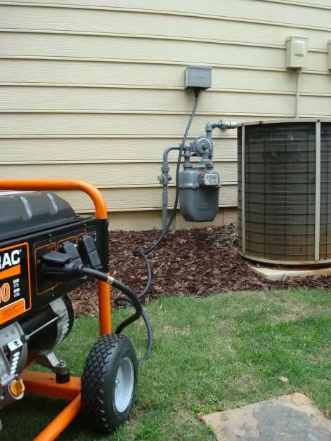 A portable generator is connected to a transfer switch on the exterior wall of a house near an air conditioning unit and gas meter.