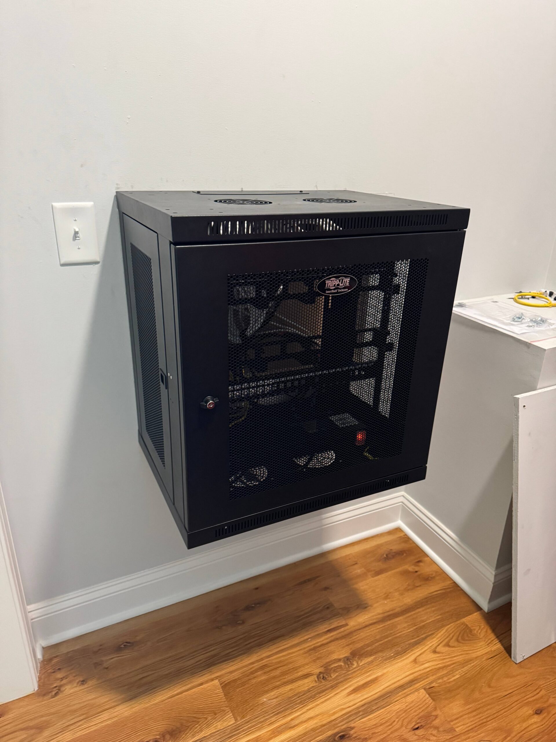 A black metal wall-mounted server rack with mesh doors is installed on a white wall above a hardwood floor.