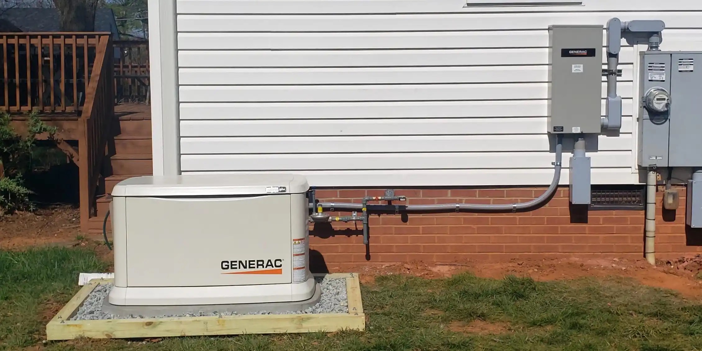 A Generac standby generator is installed next to a house, connected by metal conduit to an electrical panel mounted on a brick and siding exterior wall.