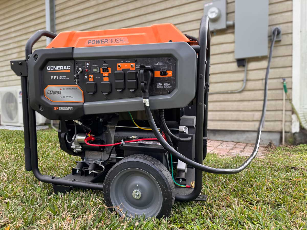 Portable Generac generator from an electric generator shop placed on grass, connected to a gray external power inlet box mounted on the side of a house.