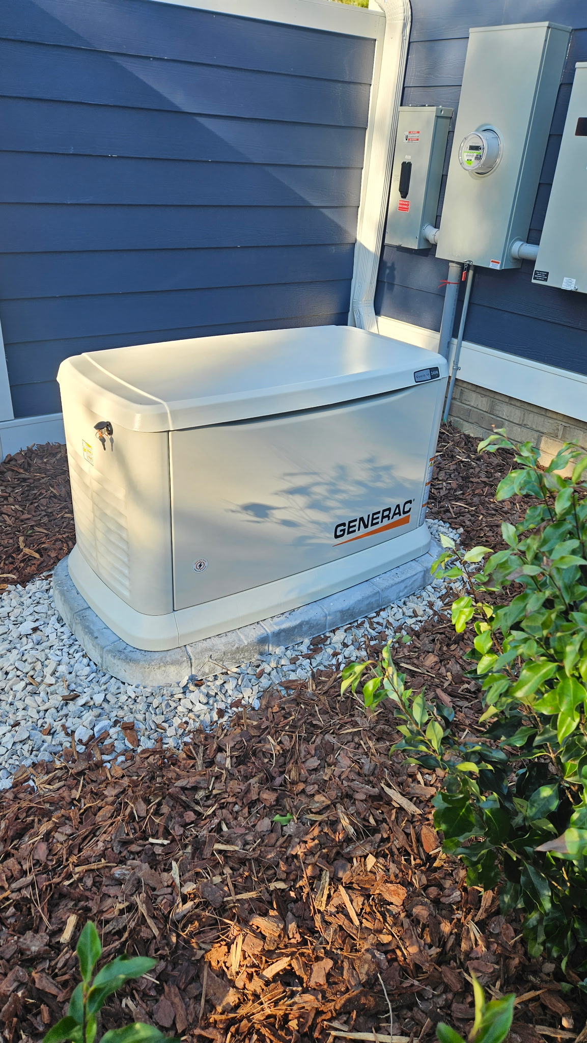 A Generac standby generator is installed on a concrete pad next to a house with blue siding, near electrical panels and mulch landscaping.