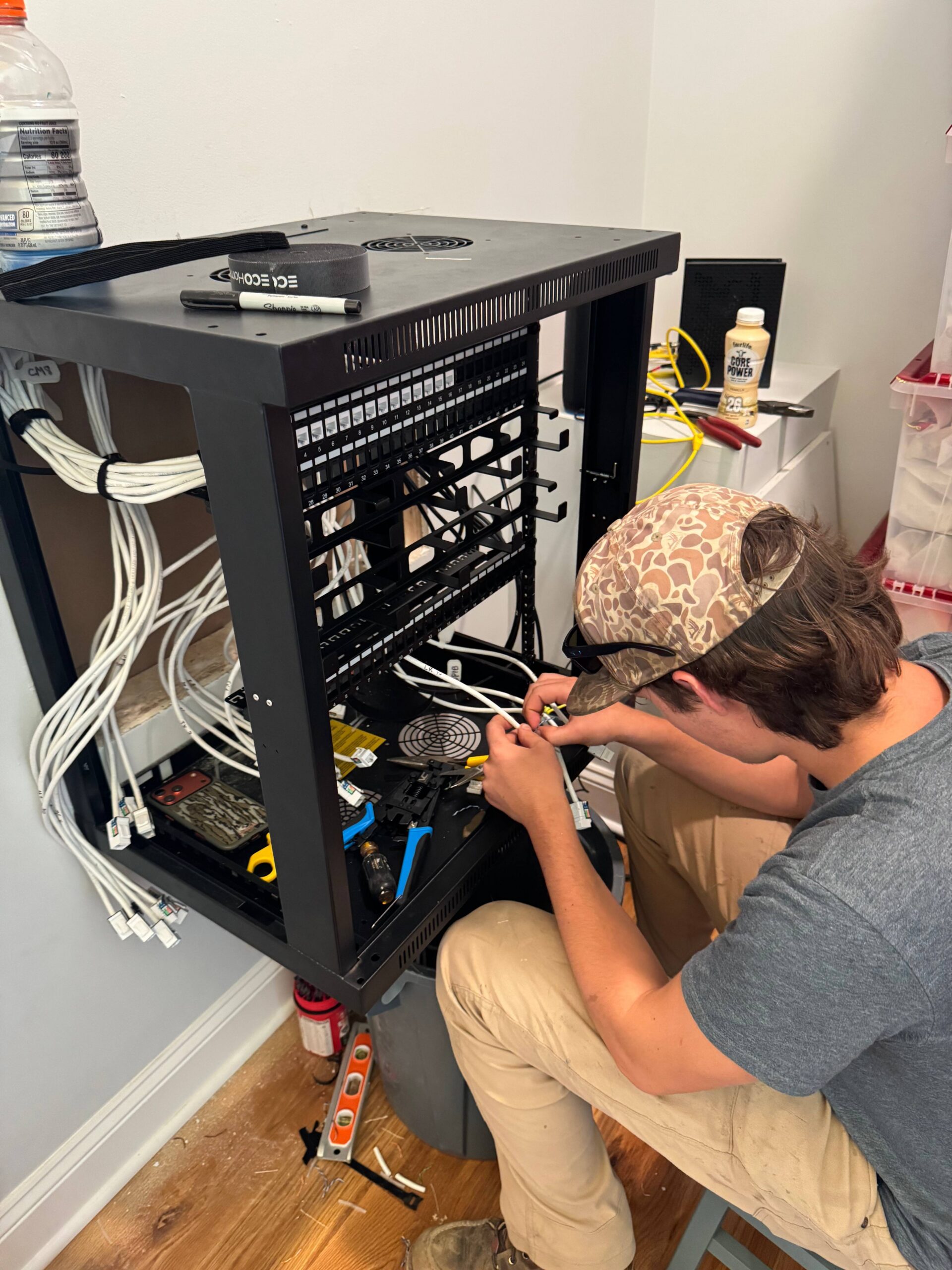 A person wearing a cap is working on network cables inside a mounted server rack, surrounded by tools and electronic equipment on a wooden floor.