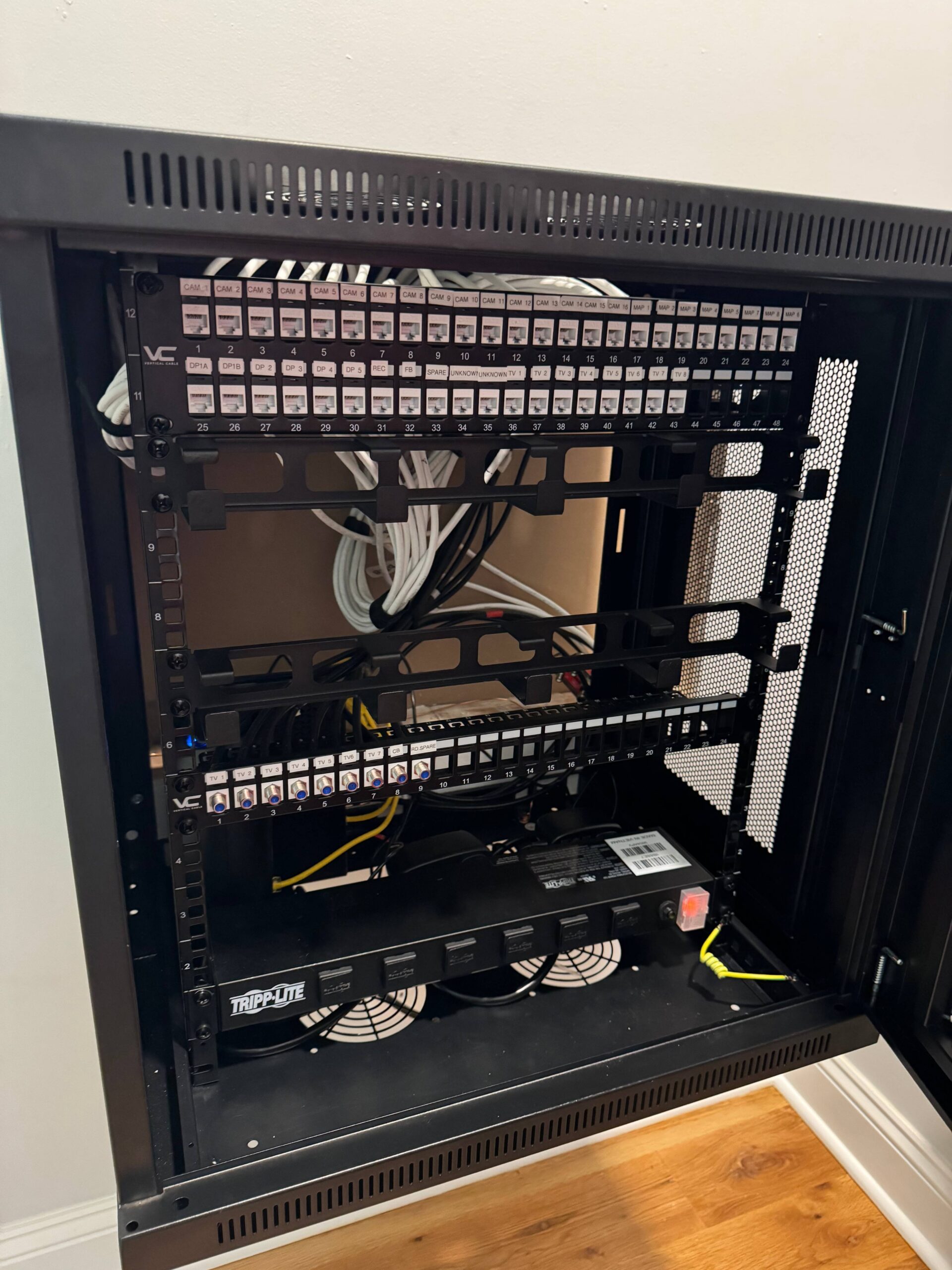 Open network rack with patch panels, organized white Ethernet cables, a Tripp Lite power distribution unit, and visible network hardware inside a cabinet against a wall.